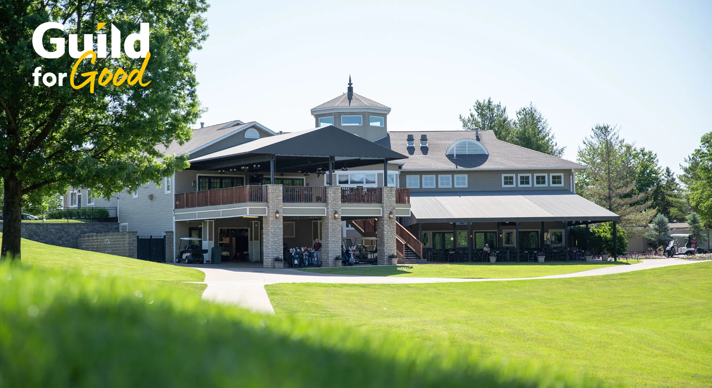 Exterior view of a golf clubhouse surrounded by green lawns and trees on a sunny day, featuring the Guild for Good logo in the upper left corner.